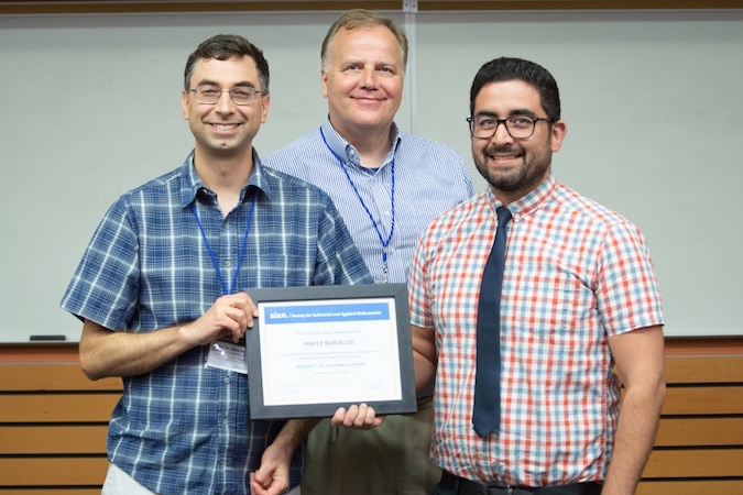 Three men facing the camera pose in front of a whiteboard with one of them holding a certificate.